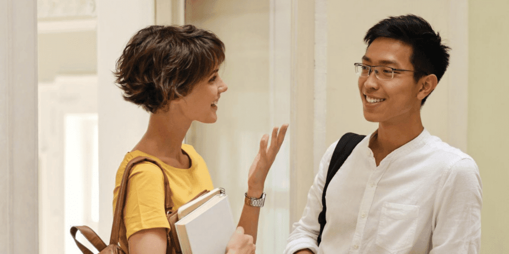 He should listen. A man listens and smiles while a woman holding a stack of books speaks to him.