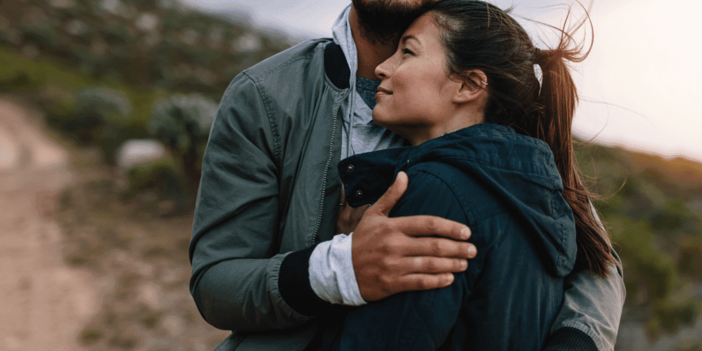 How to become more emotionally available. A man and woman embrace on a trail overlooking a sunset at the ocean.