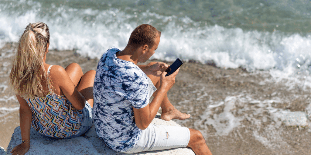 Signs your man is afraid of commitment. A man in a tie-dye t-shirt looks at his phone ignoring his girlfriend while at the beach.