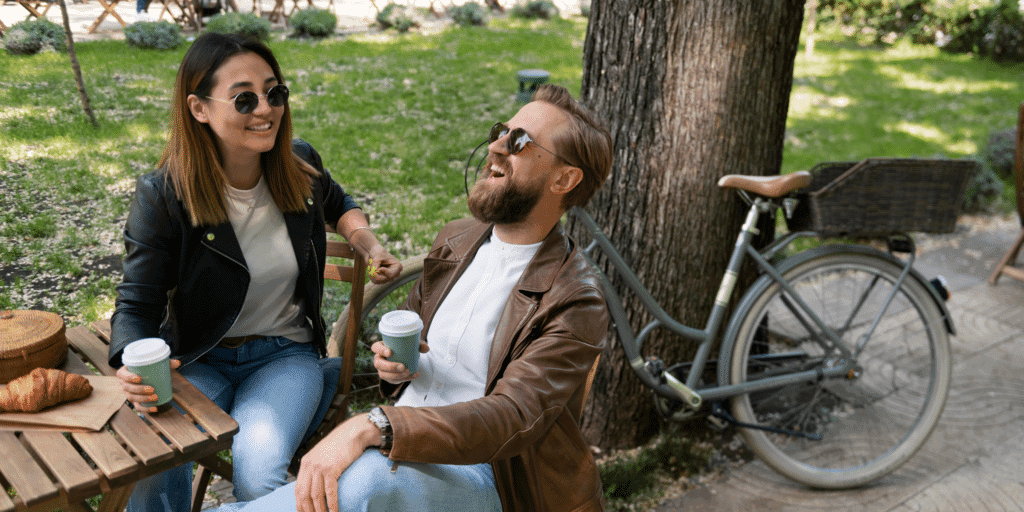A couple drinking coffee at a park table laugh together to show the importance of keeping the conversation going when you approach a woman.