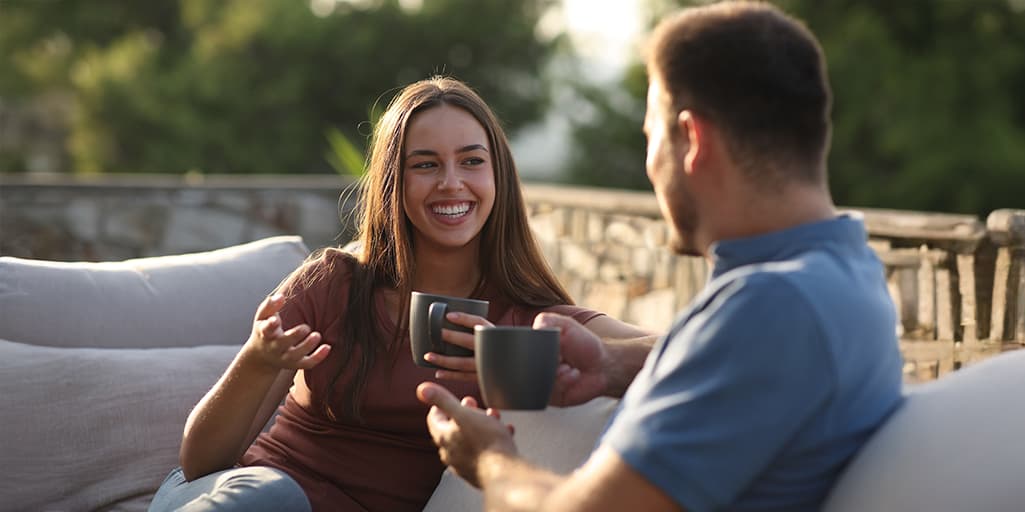 A couple happily talk over coffee on comfy patio furniture in the morning sun after meeting through a matchmaker.