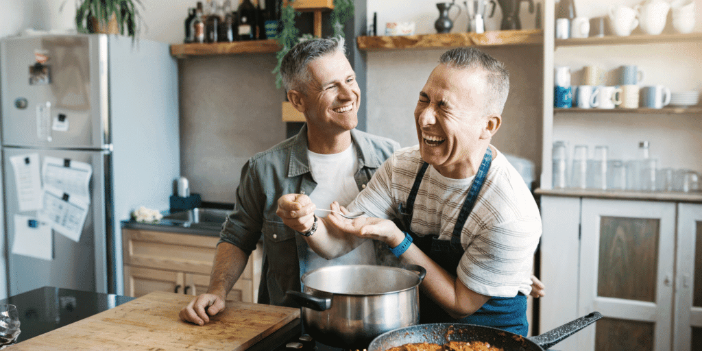 A gay male couple laugh together while they cook in the kitchen.