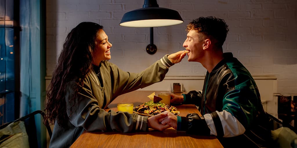 A couple has a fun moment on their date as the woman feeds the man nachos while sitting at a dining table in an apartment.
