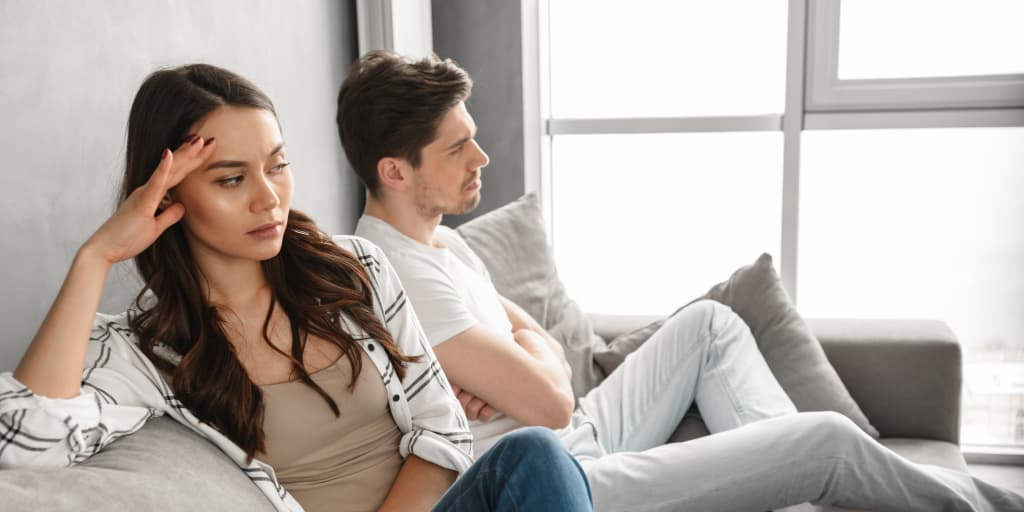 Couple sitting apart on a couch looking frustrated and distant, illustrating relationship conflict and lack of communication.